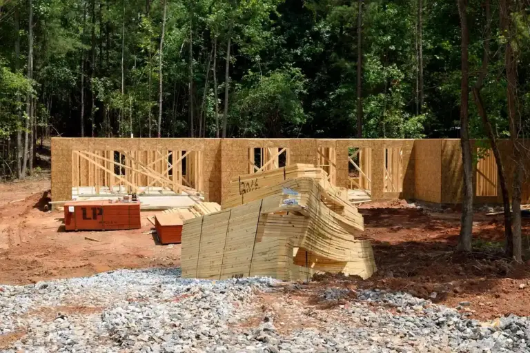 Wood-framed building under construction in a forested area with stacks of Baltic birch plywood sheets in the foreground.