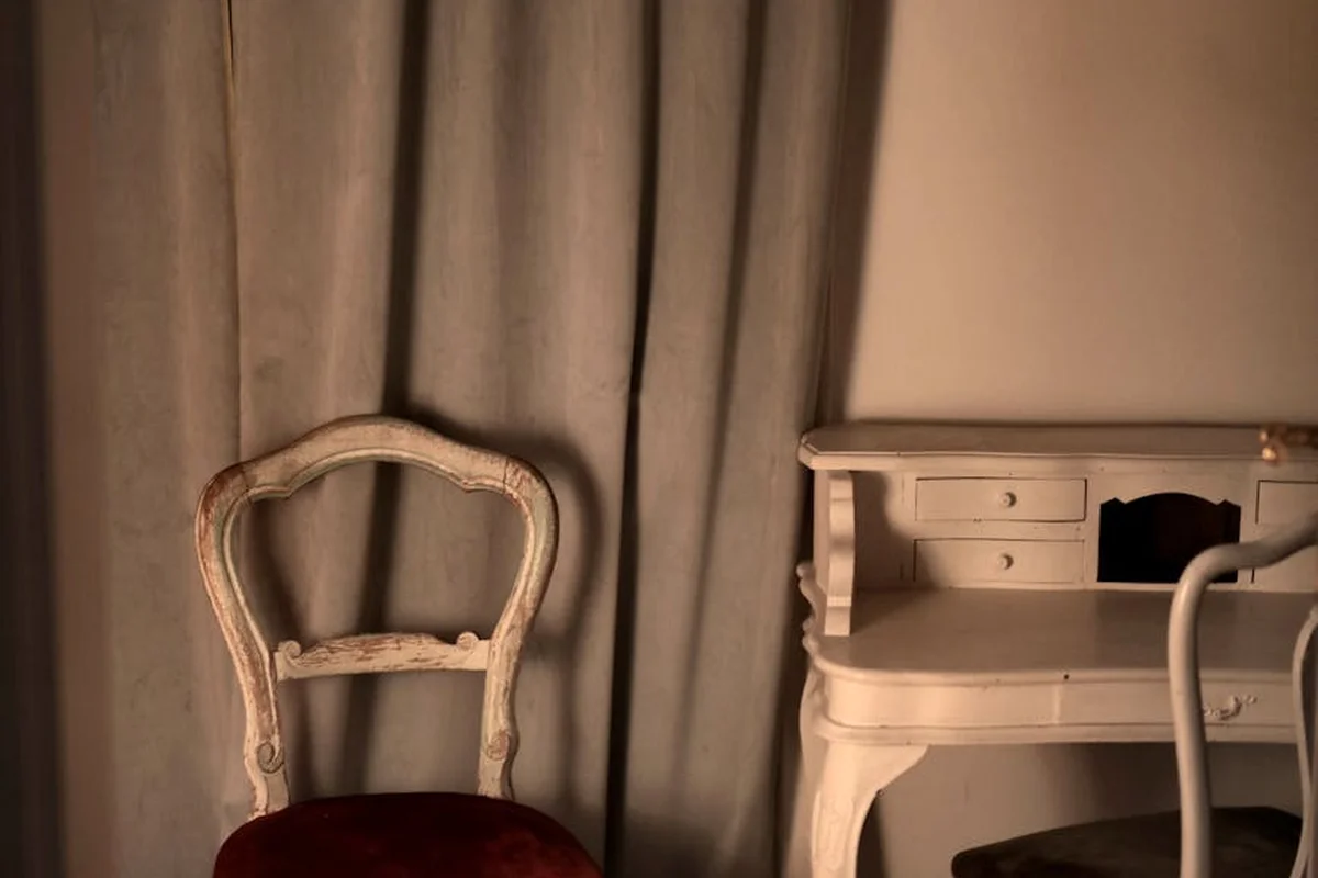 Distressed antique furniture: a chipped wooden chair sits beside a small writing desk in a softly lit room.