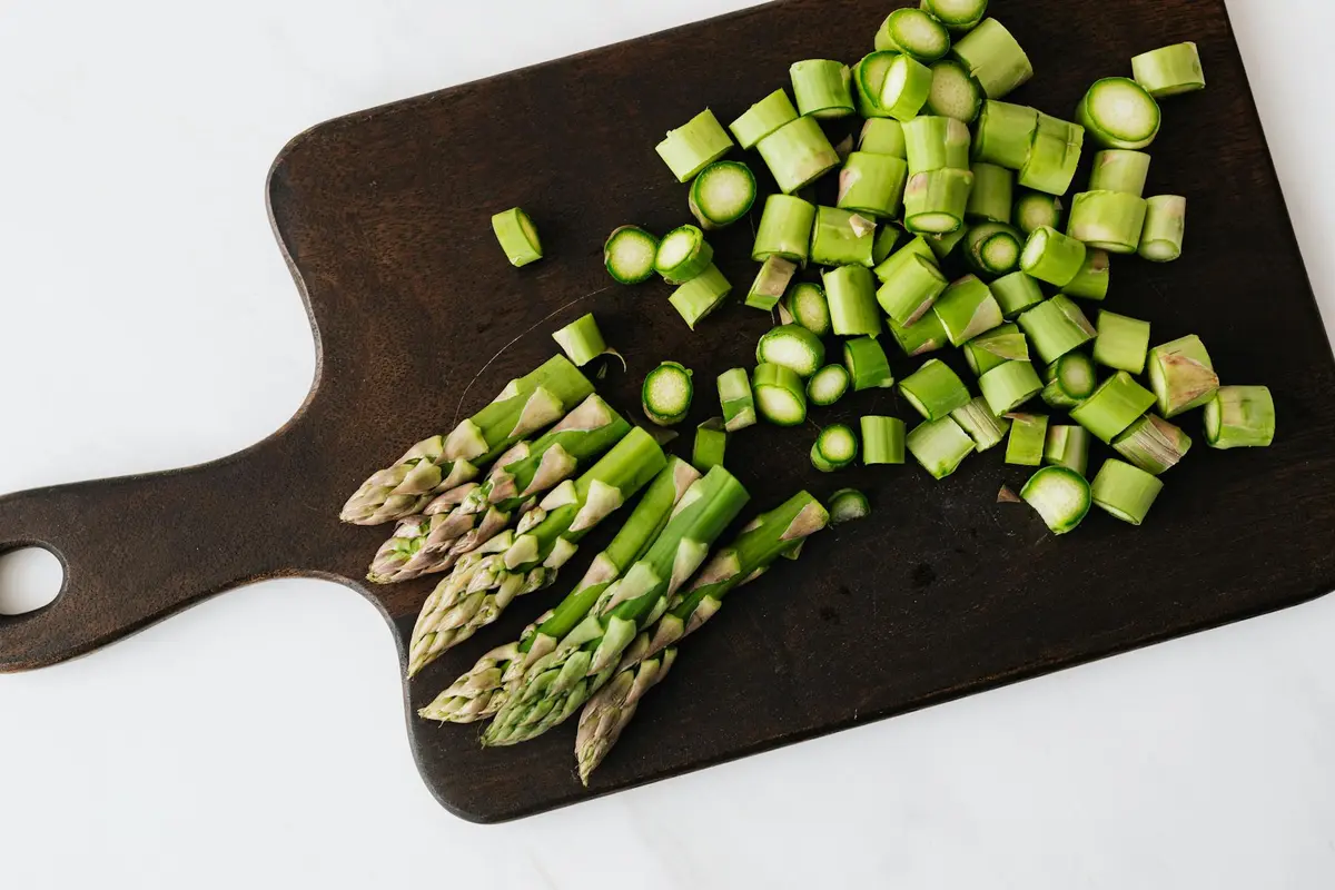 Fresh asparagus chopped into pieces on a dark wooden cutting board with a handle against a white background