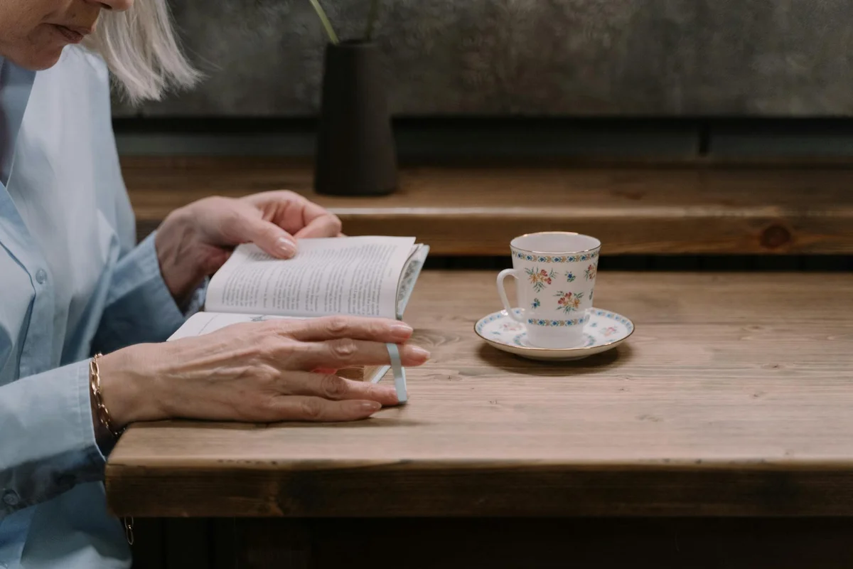 An elderly woman in a light blue shirt reads a small book at a wooden table; a delicate floral teacup and saucer sit nearby.