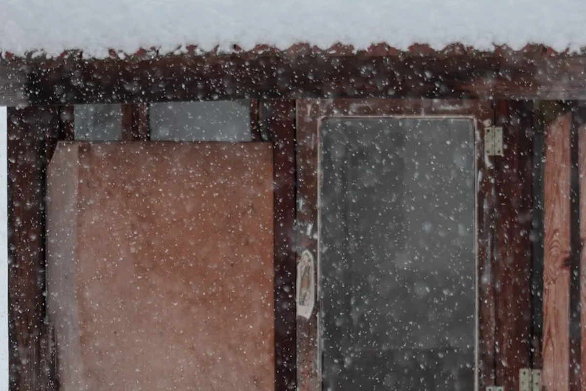Snow falling on a wooden exterior with two window openings, showing the grain and texture of the wood.