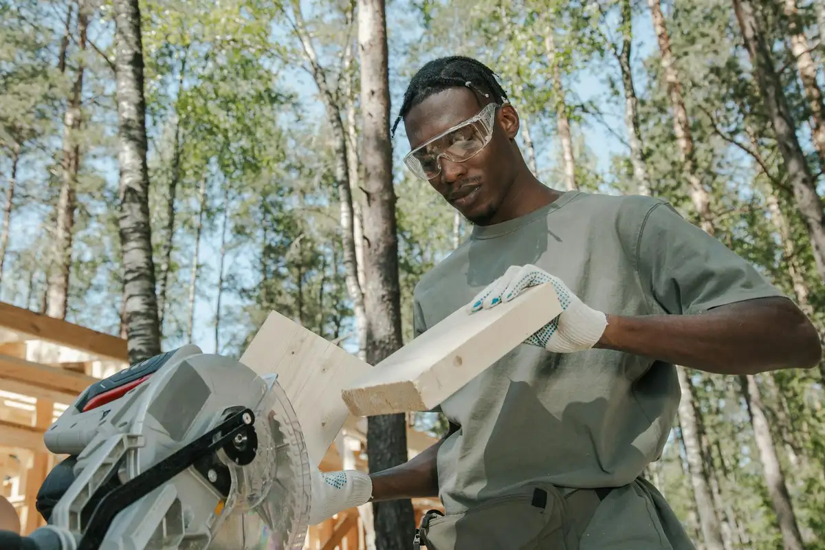 A woodworker wearing safety glasses and gloves operates a miter saw outdoors, cutting a wooden board with a blade guard visible.