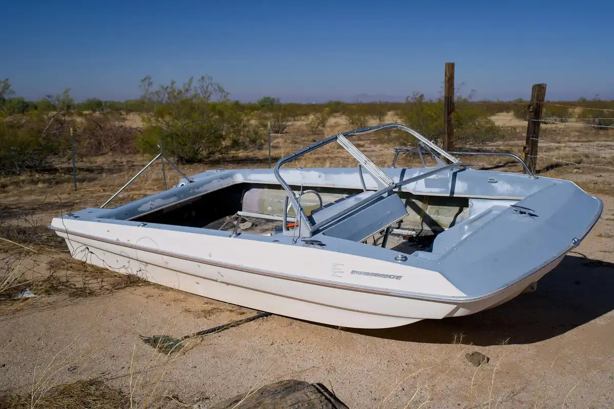 Abandoned white motorboat on sandy desert ground with sparse vegetation and a fence in the background.