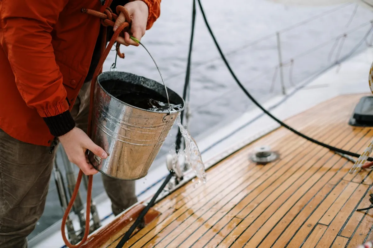 A person in an orange jacket pours adhesive from a metal bucket onto a wooden boat deck, with rigging and metal fittings visible nearby.