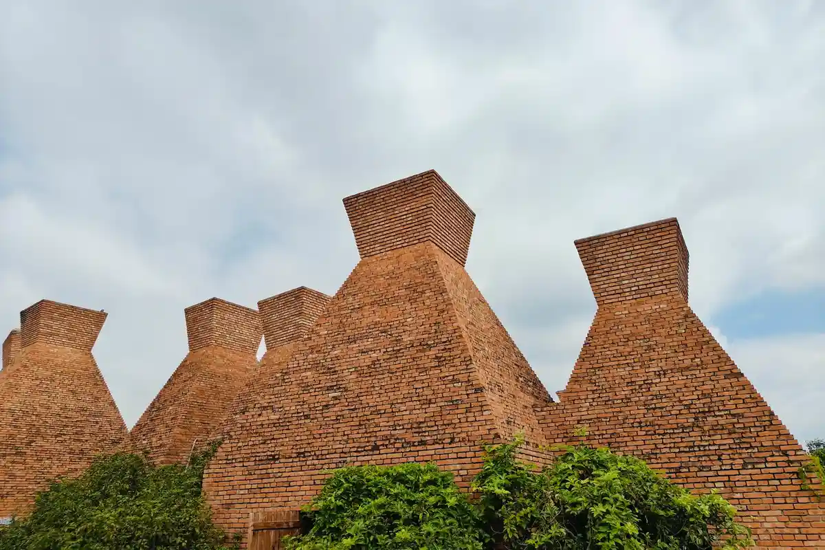 Cluster of large brick kilns with chimneys outdoors, surrounded by greenery and a cloudy sky
