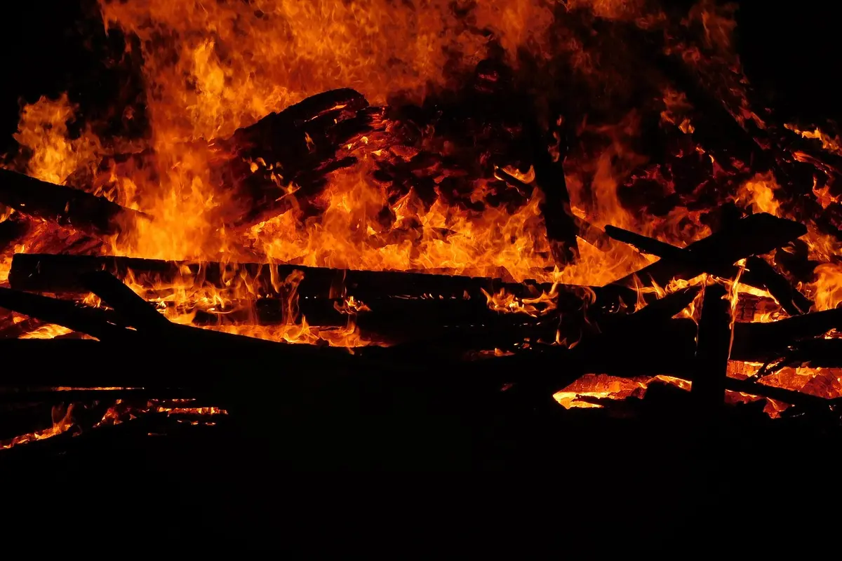 A intense blaze consumes scattered wooden boards, with bright orange flames and dark smoke rising into the air.