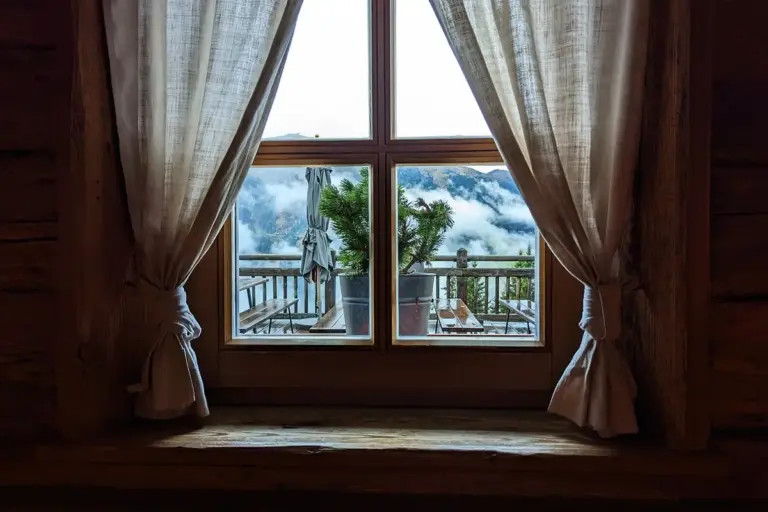 A cozy cabin window with light curtains framing a view of a wooden balcony and cloudy sky outside.