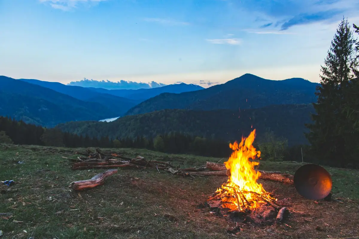 Campfire burning in a grassy meadow with a distant mountain range, illustrating outdoor wood smoking and flavor comparison for hickory.