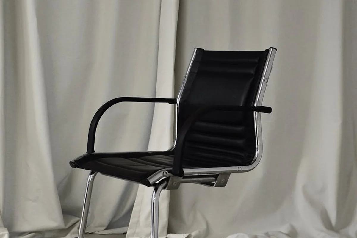 A black leather and metal office chair sits against a light, draped backdrop.