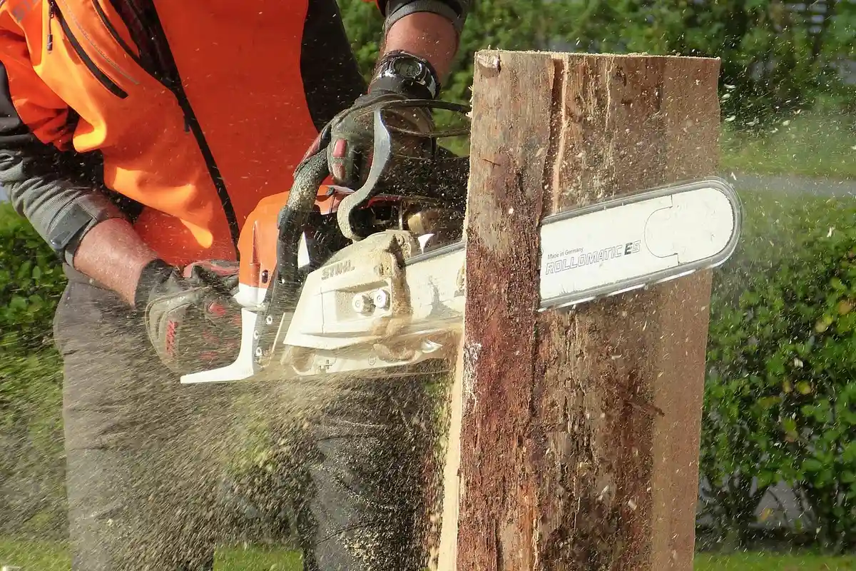 Worker wearing an orange safety vest and gloves operating a power saw to cut a vertical log outdoors, with wood dust visible in the air.