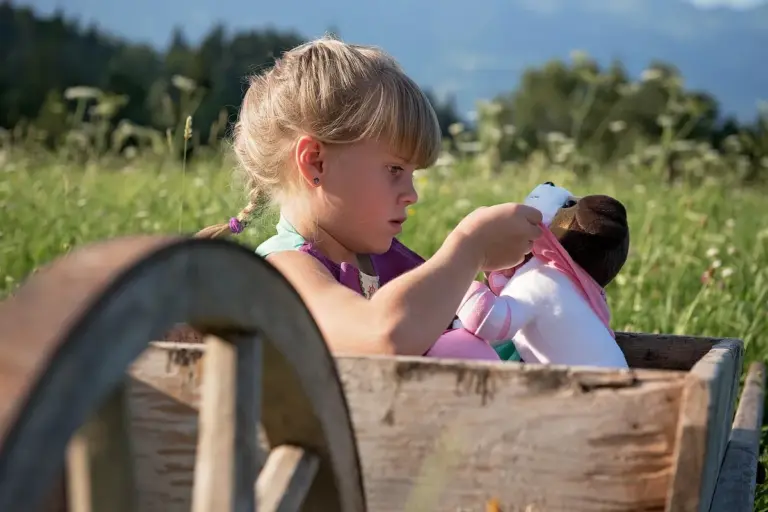 A young girl sits in a rustic wooden wagon outdoors, playing with a doll.