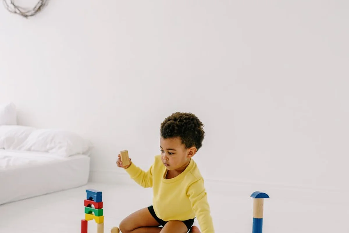 A toddler sits on a bright, white floor in a minimalist room, stacking colorful wooden blocks.