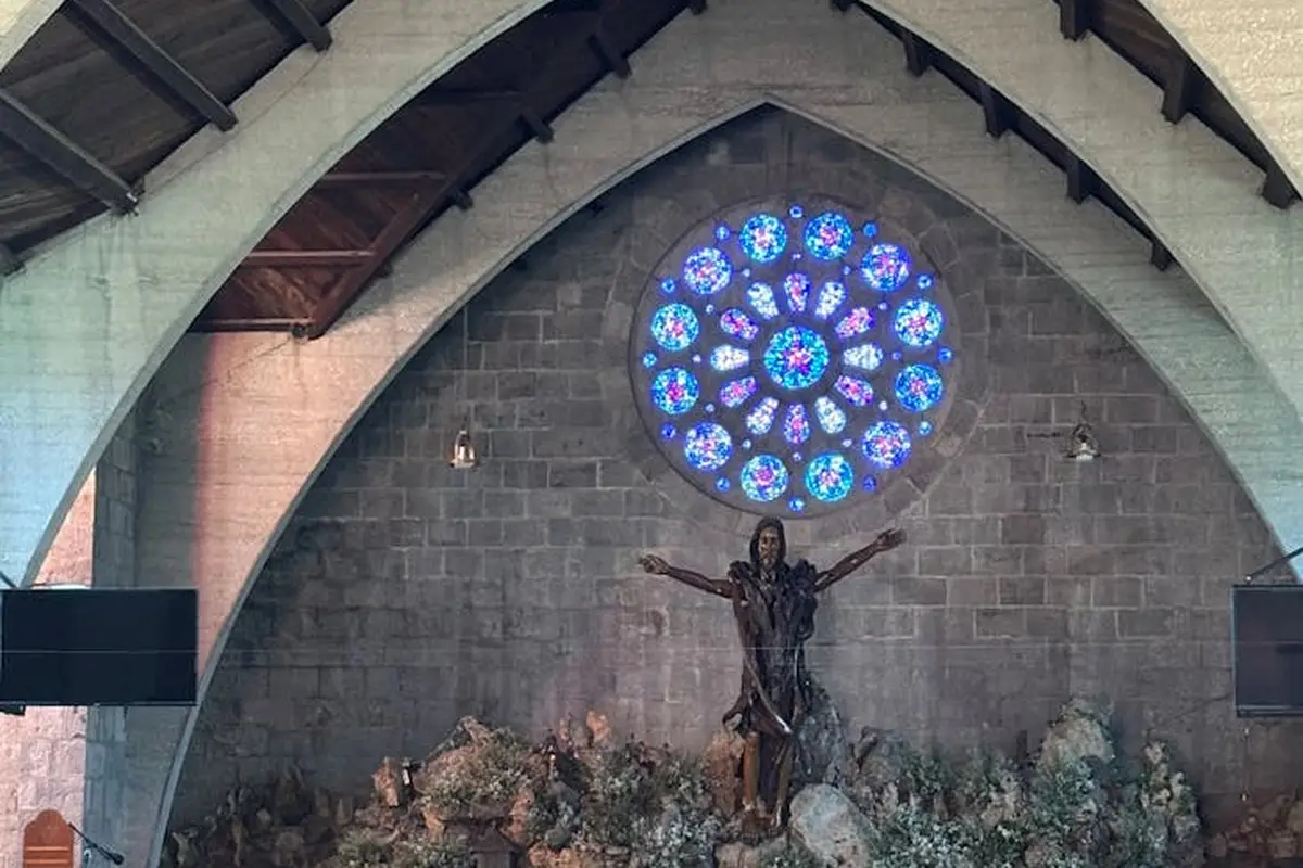 Interior of a church featuring a circular stained-glass rose window above an altar statue, with stone arches and floral arrangements.