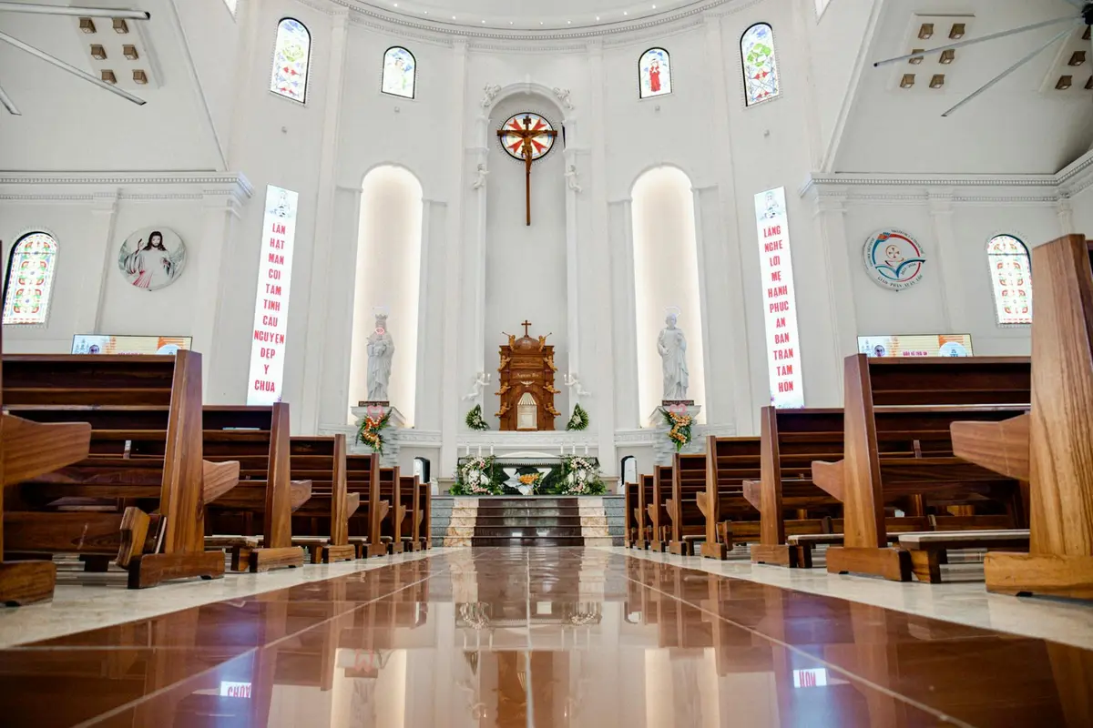 Interior view of a church with wooden pews, an ornate altar, and tall arched windows allowing light to fill the nave.