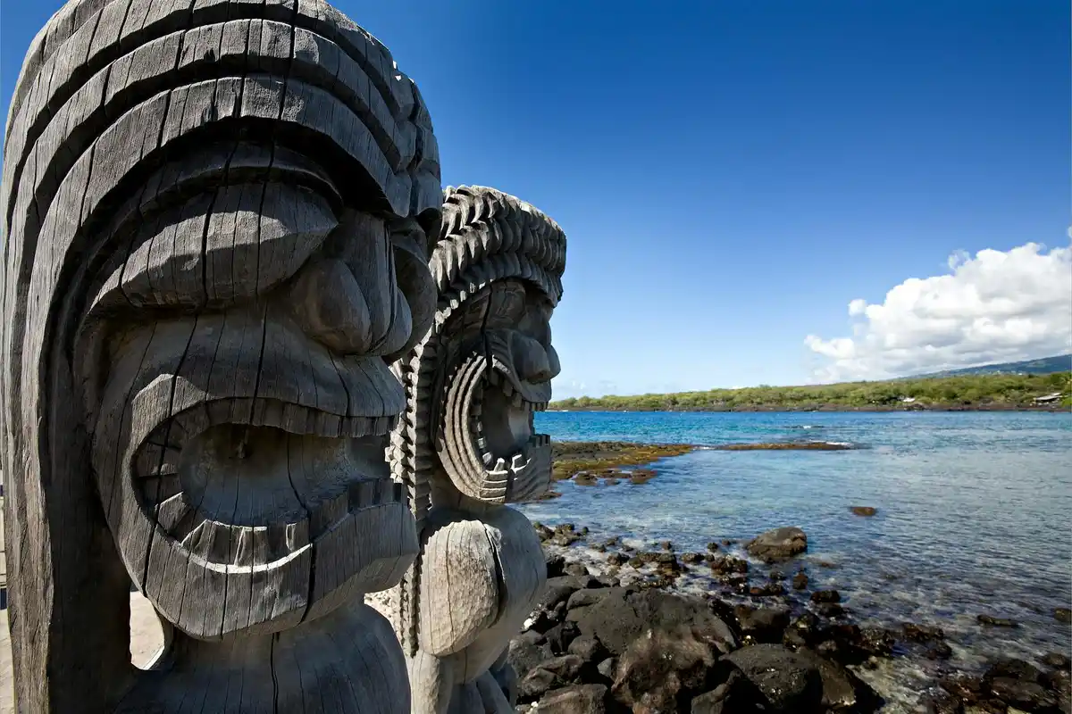 Carved wooden totem sculptures along a rocky shoreline under a bright blue sky.