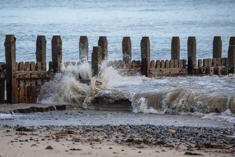 Waves crash against a wooden breakwater along a shore, spraying water over the posts.