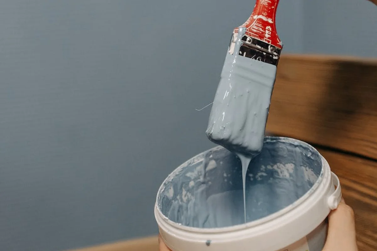 Close-up of a paintbrush loaded with gray paint dipping into a can and applying paint to a wooden surface.