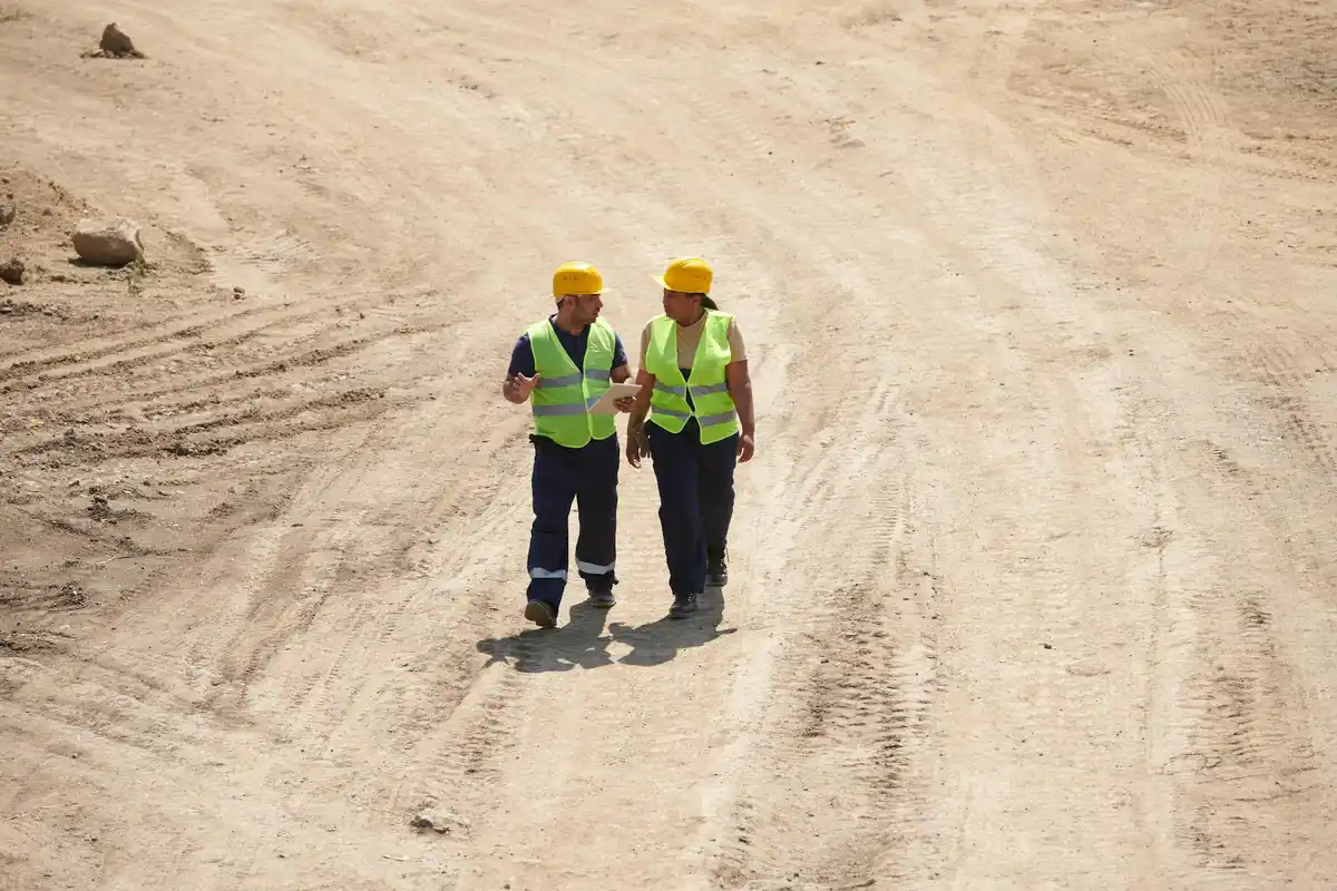 Two construction workers wearing yellow hard hats and high-visibility vests standing on a dusty construction site.