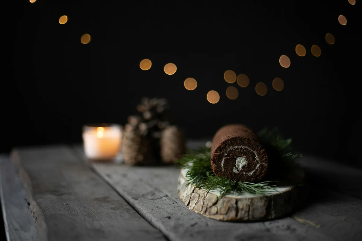 A rustic scene featuring a cake placed on a wooden log slice atop a weathered wooden table, with candlelight and warm bokeh in the background.