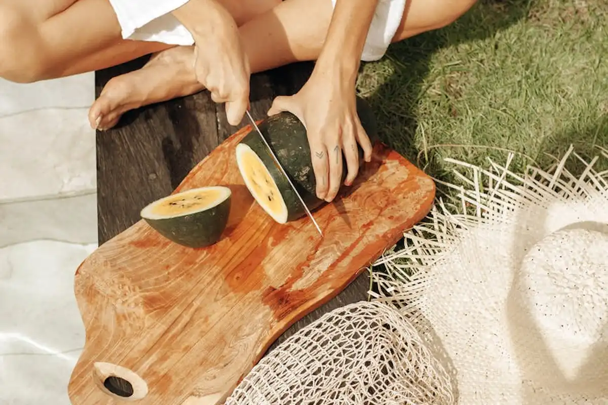 Two hands slice a green-skinned fruit on a wooden cutting board outdoors, with a mesh bag and white towel nearby.