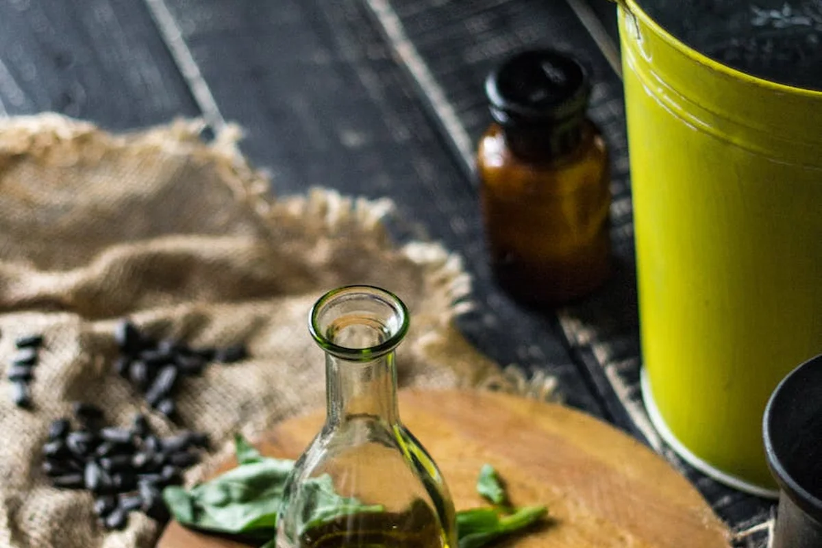 Close-up of a glass oil bottle on a wooden work surface with burlap, leaves, and a yellow container in the background.