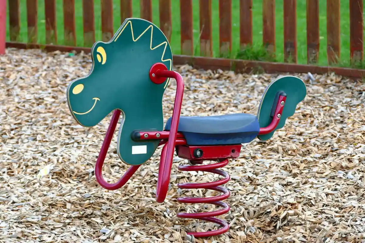 Green dinosaur-shaped playground spring rider on a wood-chip surface with a red frame and blue seat, set against a wooden fence.