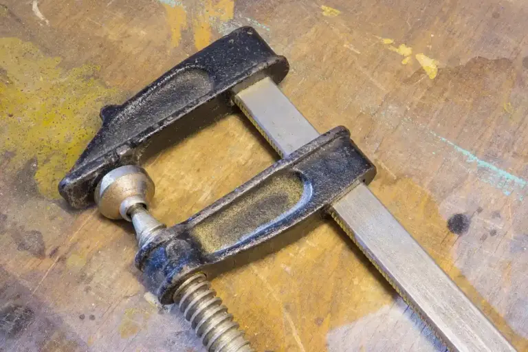 A metal C-clamp clamps a metal bar on a worn wooden workbench