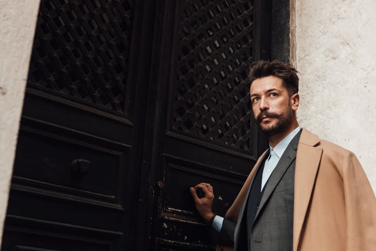 A man in a tan overcoat and suit stands beside a dark wooden door with weathered panels, suggesting inspection of door joints and wood condition.