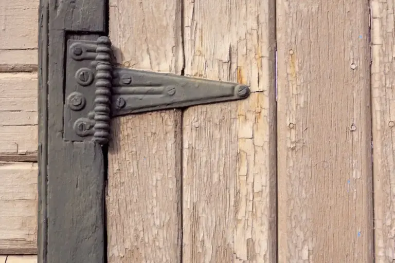 Close-up of a weathered wooden door with a metal hinge, showing cracks and wear around a loose joint.