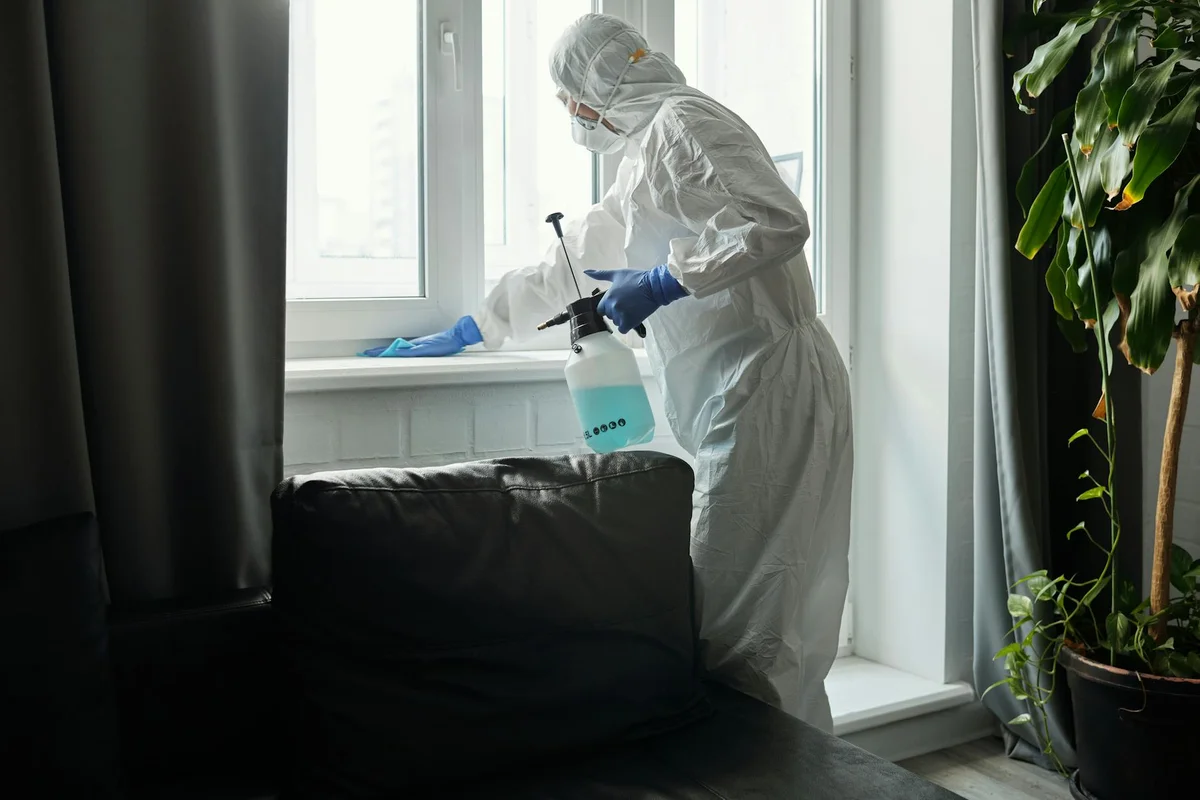 A person in full protective PPE uses a spray bottle to clean near a window in a living room, with a dark sofa and houseplants nearby.