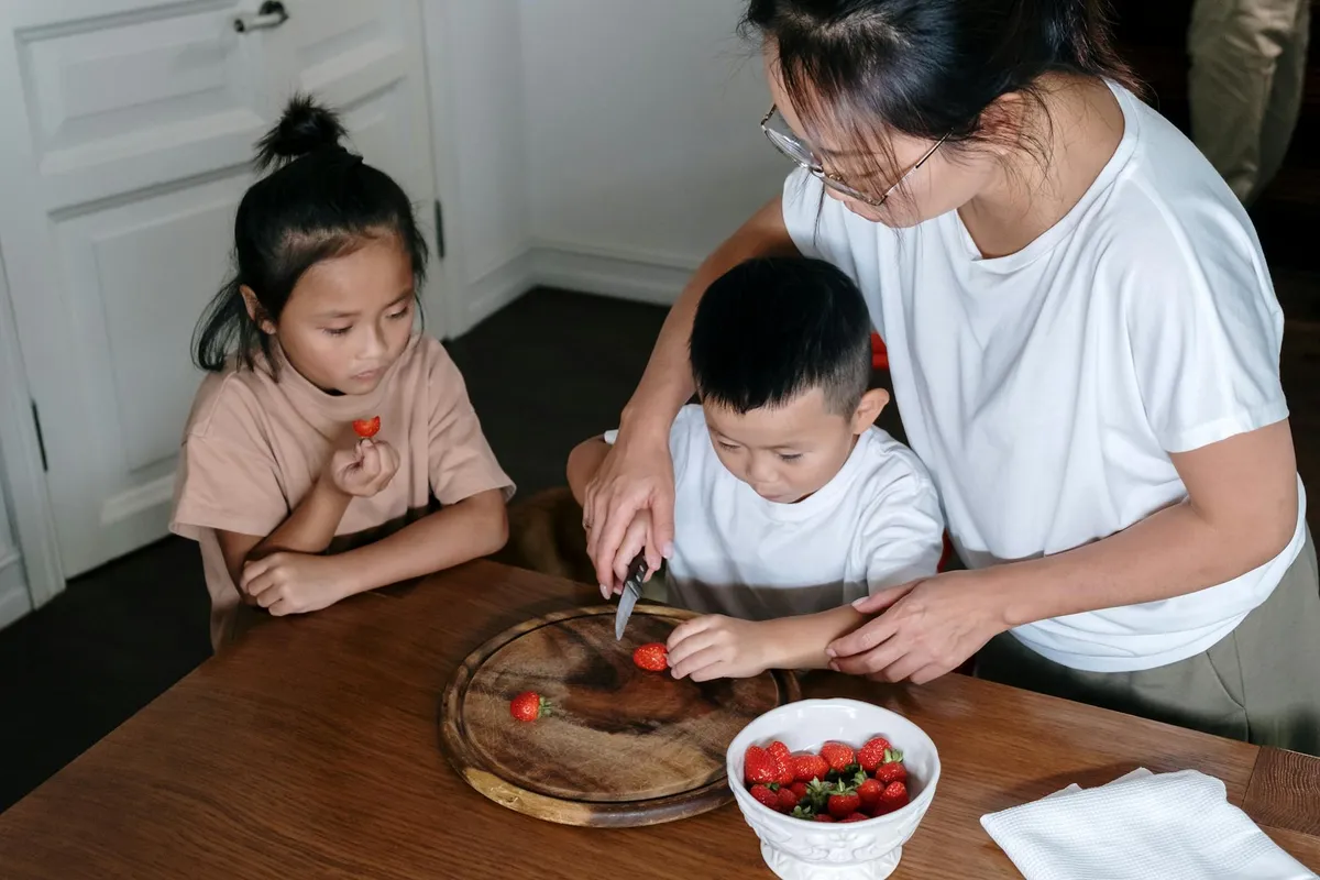 Mother and two children cutting strawberries on a wooden cutting board at a kitchen table.