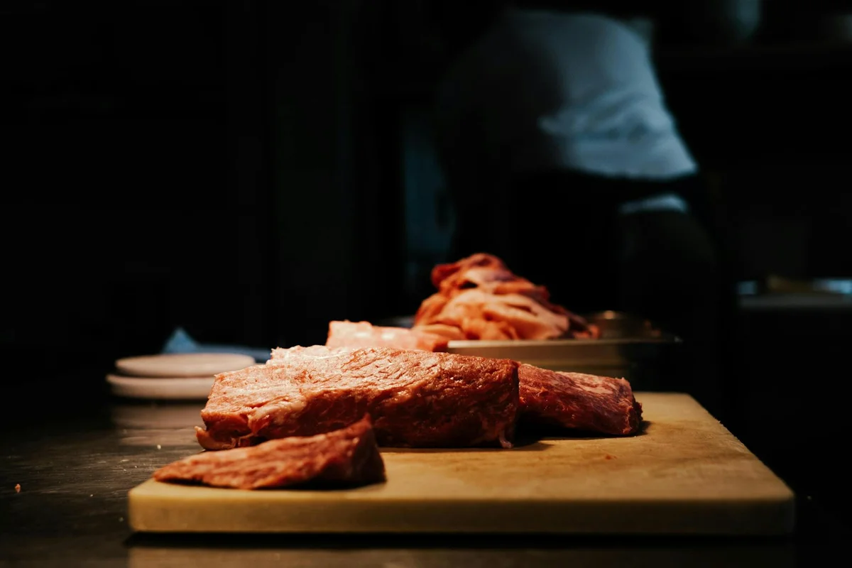 Close-up of raw meat on a wooden cutting board in a dim kitchen with a blurred chef in the background.