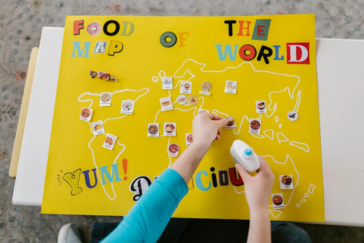 Hands applying glue to a bright yellow world-map poster with small picture stickers on it.