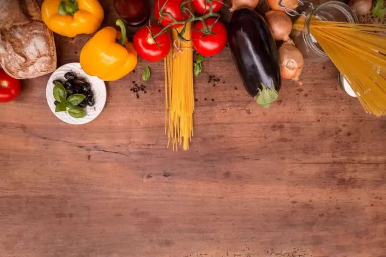 Top-down view of a wooden kitchen counter with colorful vegetables, pasta, and bread arranged around the edges.