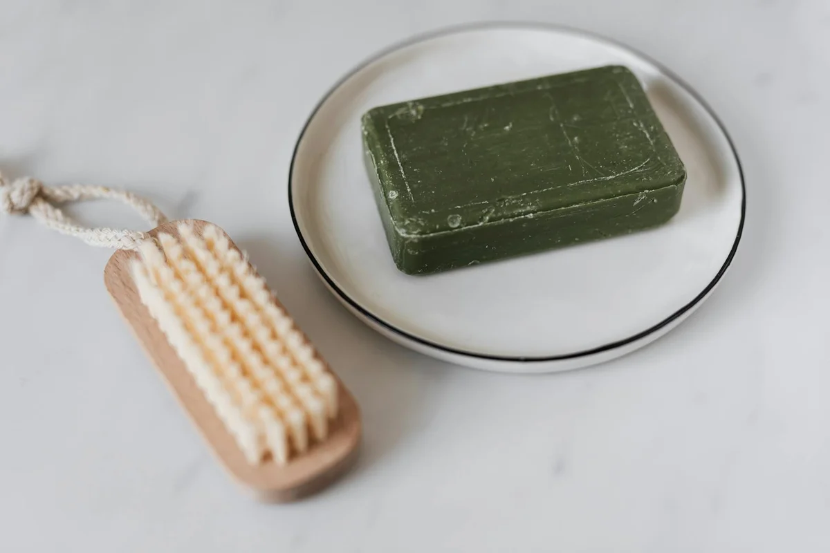 Green rectangular soap bar on a white plate beside a wooden bristle brush on a light marble surface.