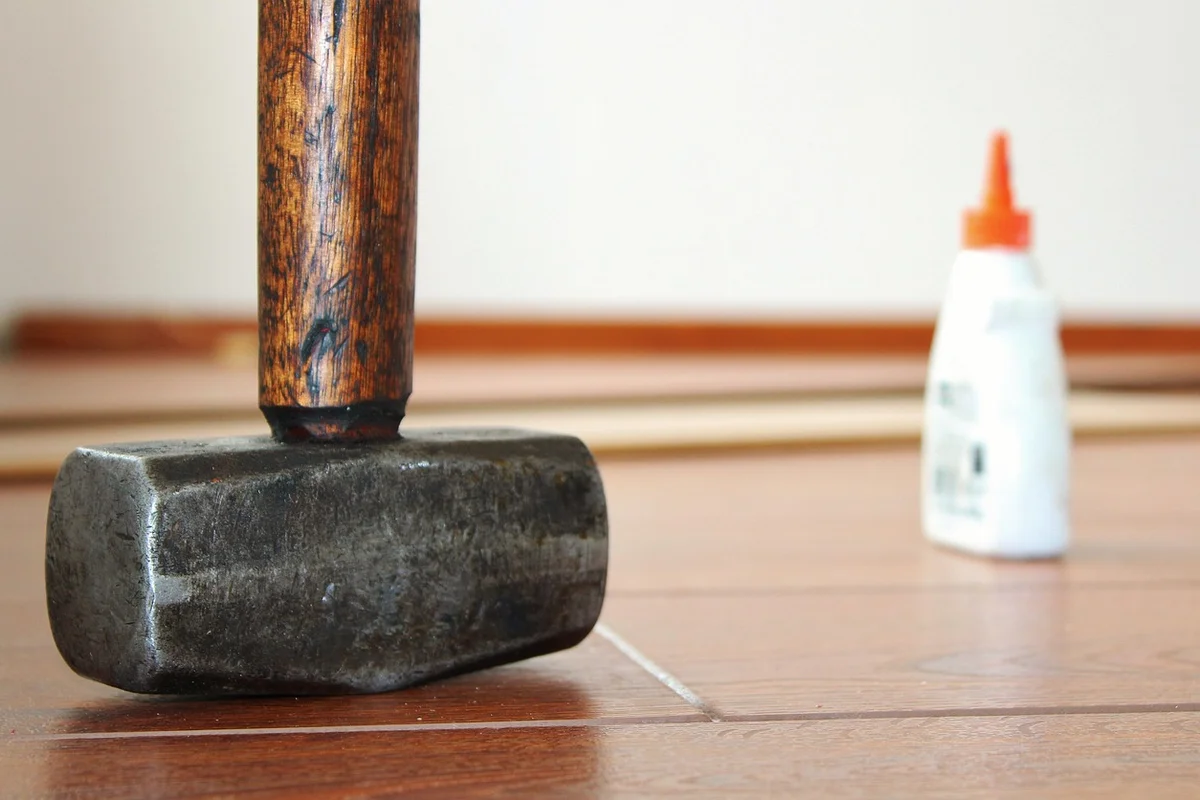 Hammer resting on a wooden floor with a bottle of glue in the background, illustrating non-wood bonding for metal, concrete, and fabric applications.