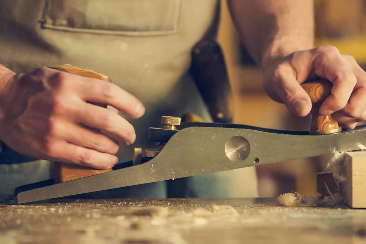 Close-up of a woodworker planing a board with a hand plane, wood shavings visible on the workbench.