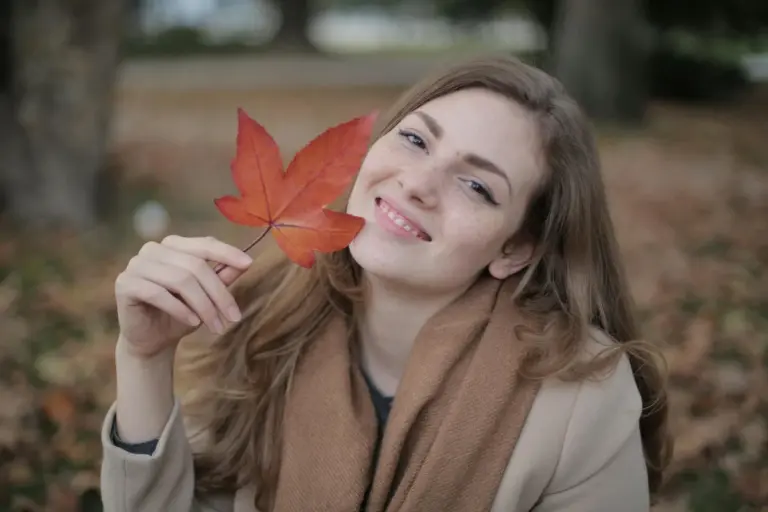 Smiling woman outdoors holding a red autumn leaf with fall colors in the background