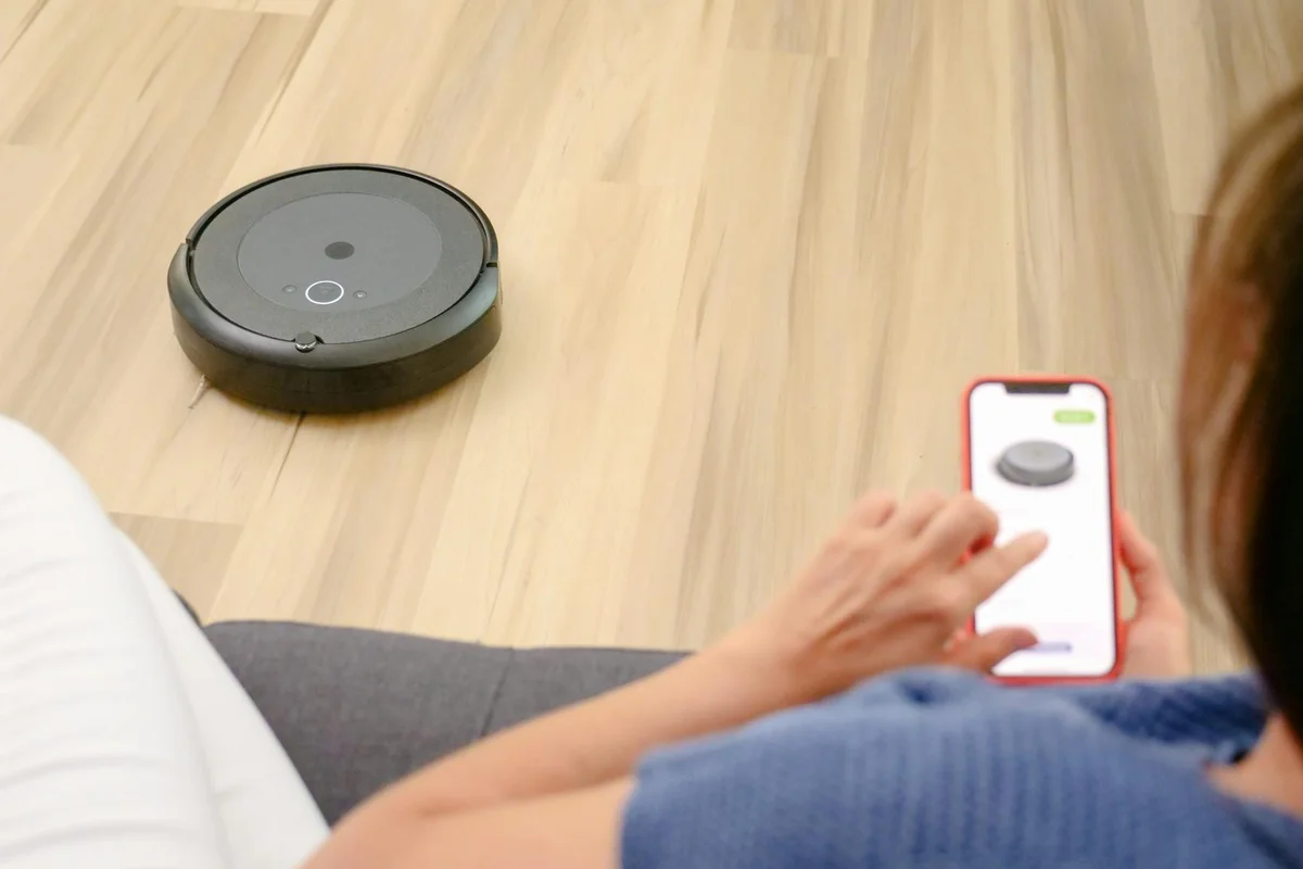 A robotic vacuum on a light hardwood floor and a person holding a smartphone to control it, illustrating the cleaning preparation step in surface restoration.