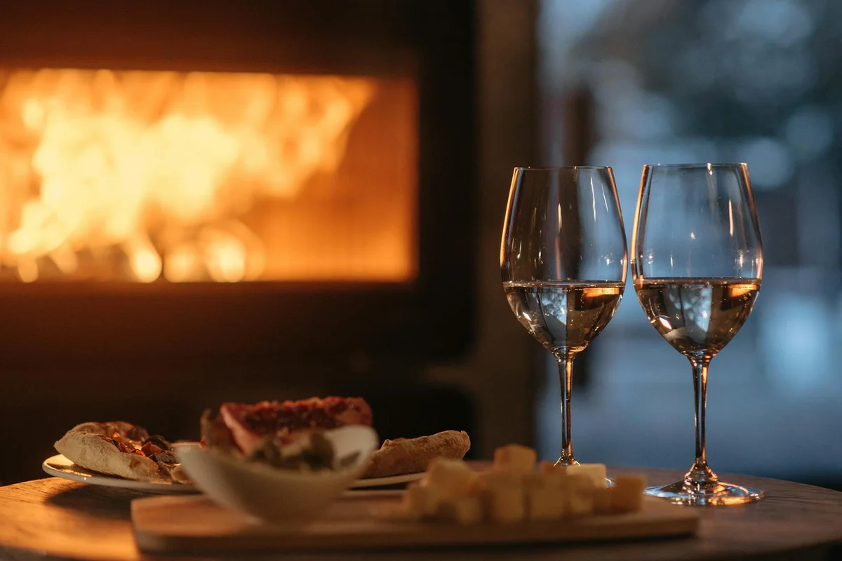 Two crystal wine glasses on a table with small plates of appetizers in front of a glowing fireplace.