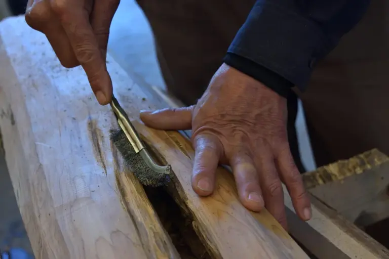 Close-up of hands using a small scraper to work on a groove in a light wood beam, exposing the grain and old finish.