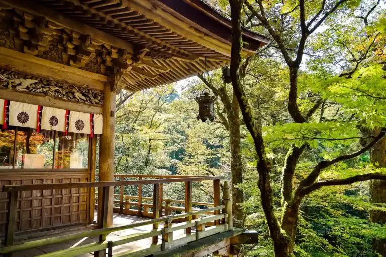 A traditional wooden shrine or temple pavilion nestled among green trees, featuring carved eaves, a wooden railing, and hanging lanterns.