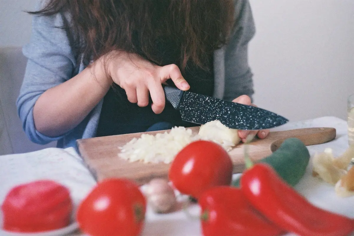 Person slicing onions on a wooden cutting board with red peppers and tomatoes nearby.