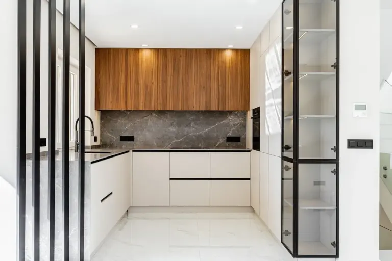 Modern L-shaped kitchen with white base cabinets, wood upper cabinets, dark marble backsplash, and glass-front pantry cabinet on the right.