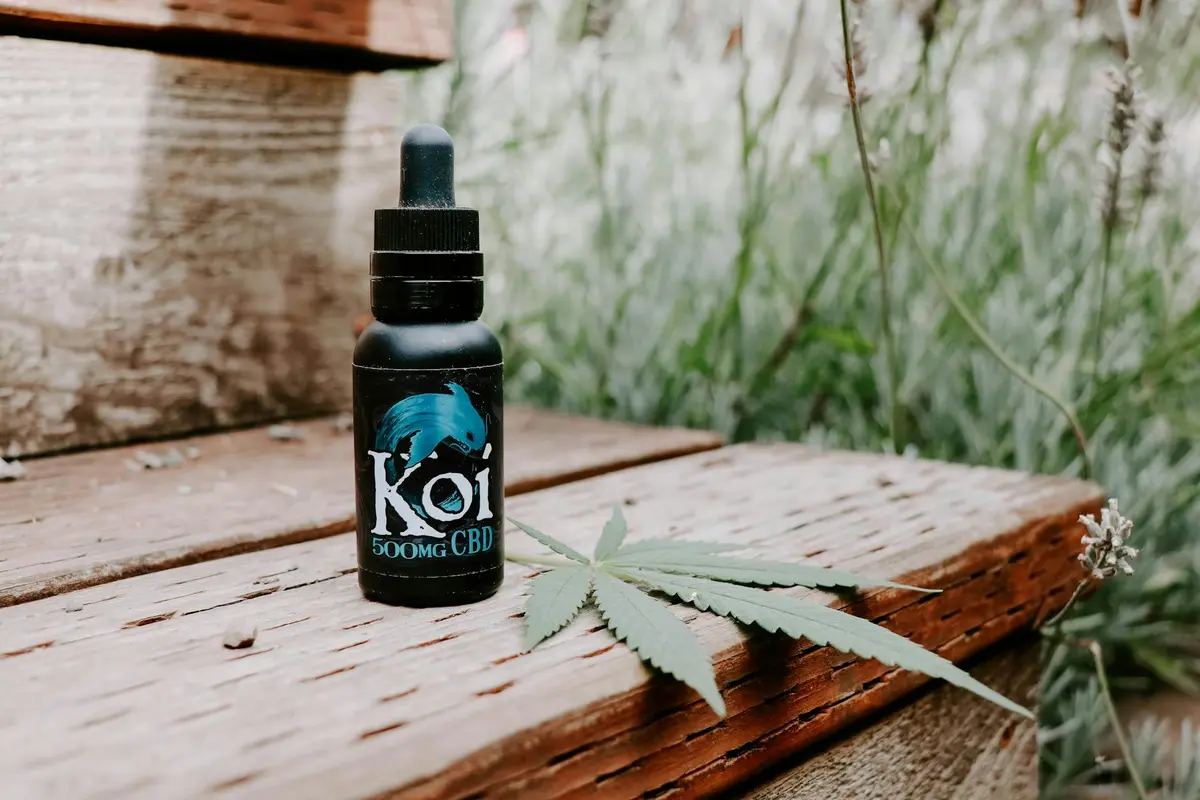 A small dark glass bottle labeled KOI CBD with a dropper sits on weathered wooden steps, with a green leaf nearby in an outdoor setting.