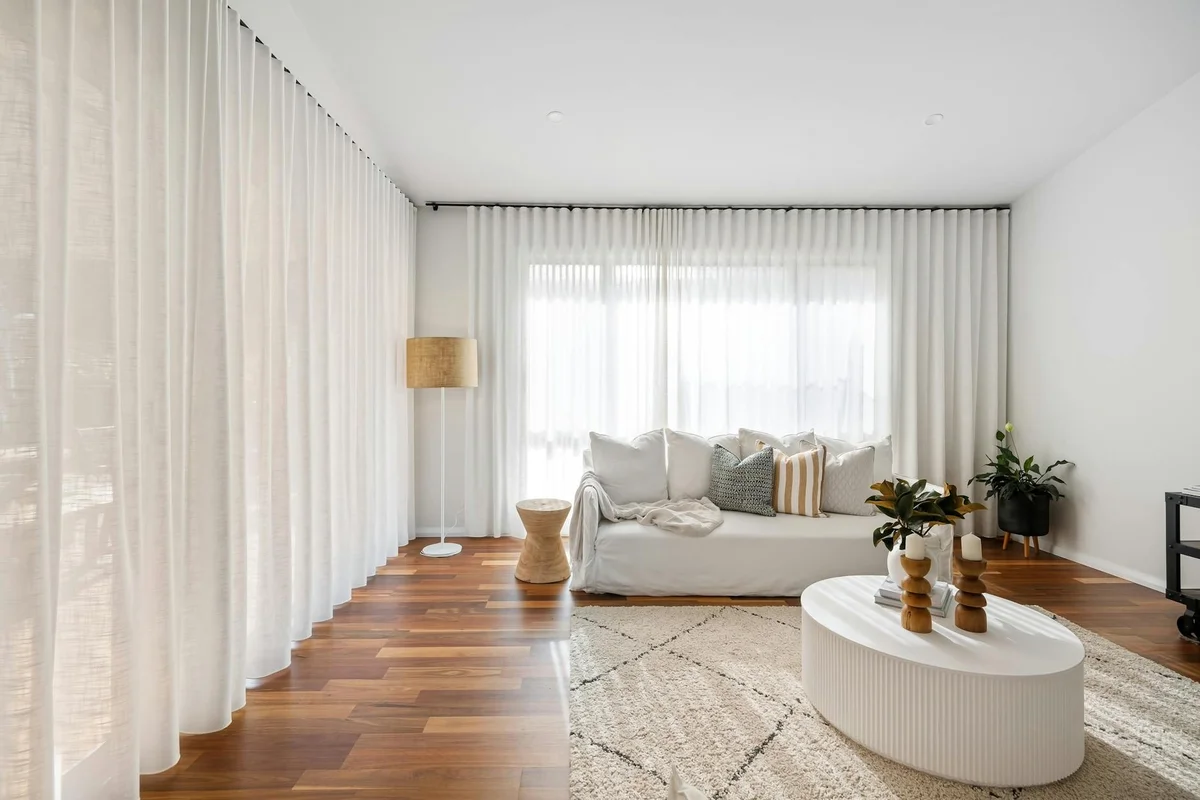 Bright minimalist living room with white walls, sheer curtains, a light gray sofa, wooden floor, and a round white coffee table.