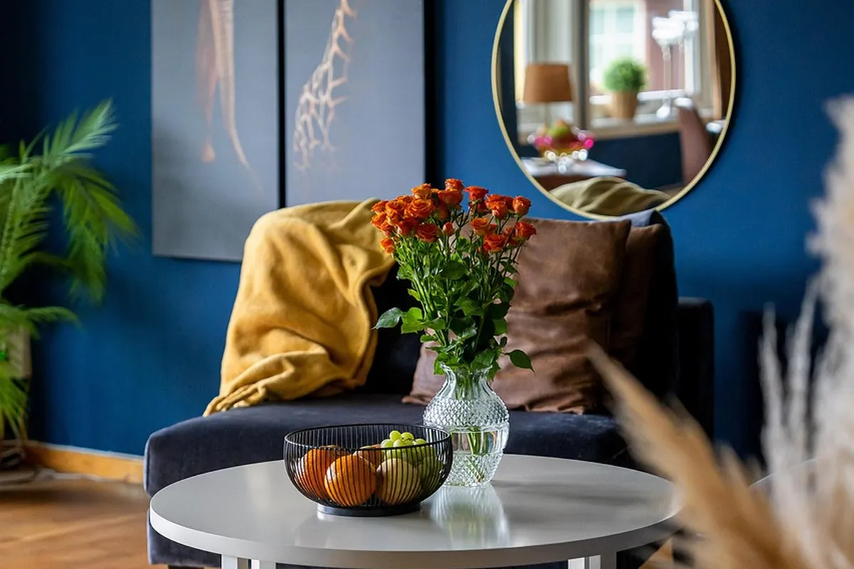 Modern living room with a blue wall, round white coffee table, a vase of orange-red flowers, a fruit bowl, a mustard throw over a dark sofa, and a round mirror reflecting a window.