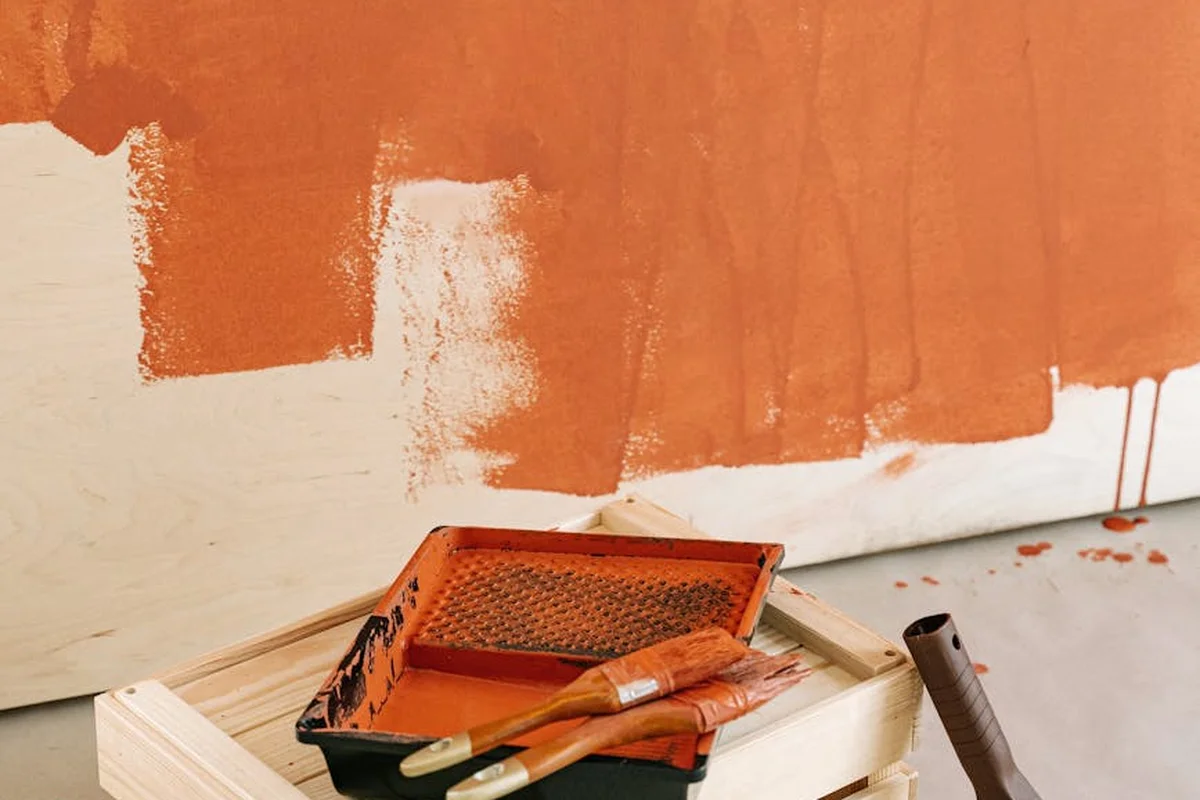 Paint tray with orange oil-based paint and brushes resting on a wooden surface beside a partially painted wall.
