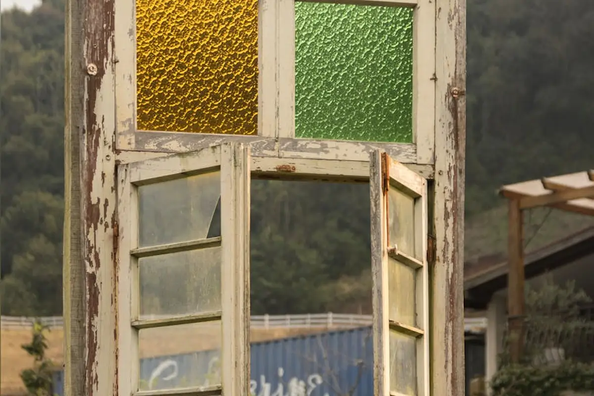 Old weathered window frame with peeling white paint and yellow and green textured stained-glass panels at the top.