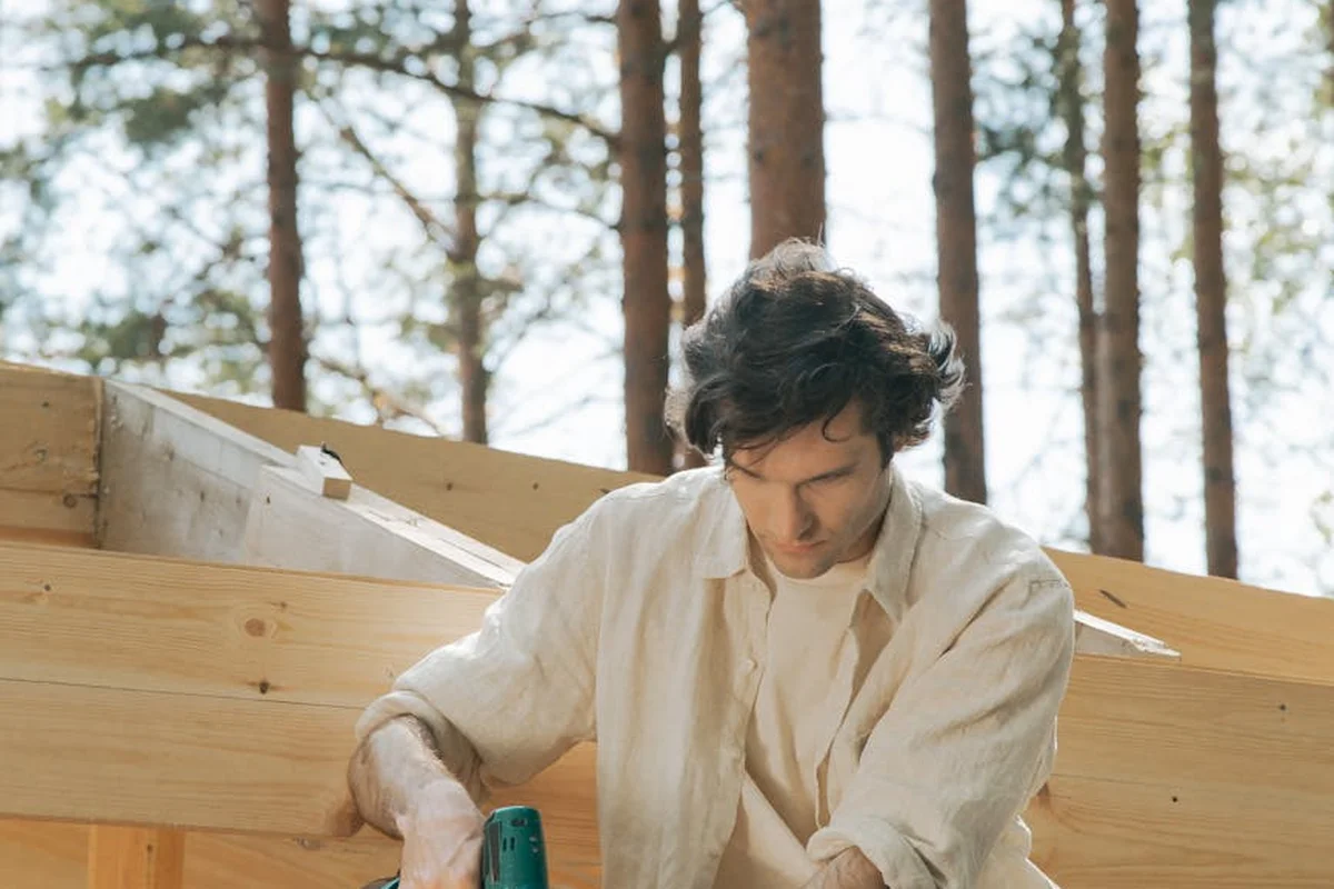 Carpenter in a light shirt working on wooden exterior framing outdoors with a power tool